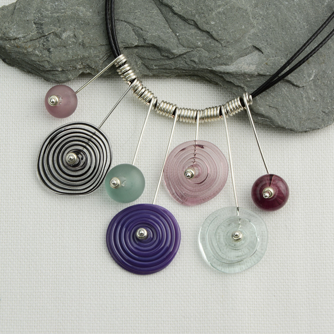 Four spiral beads and three spacer beads in shades of purple and grey on silver headpin drops with silver spiral spacers on black necklace cord