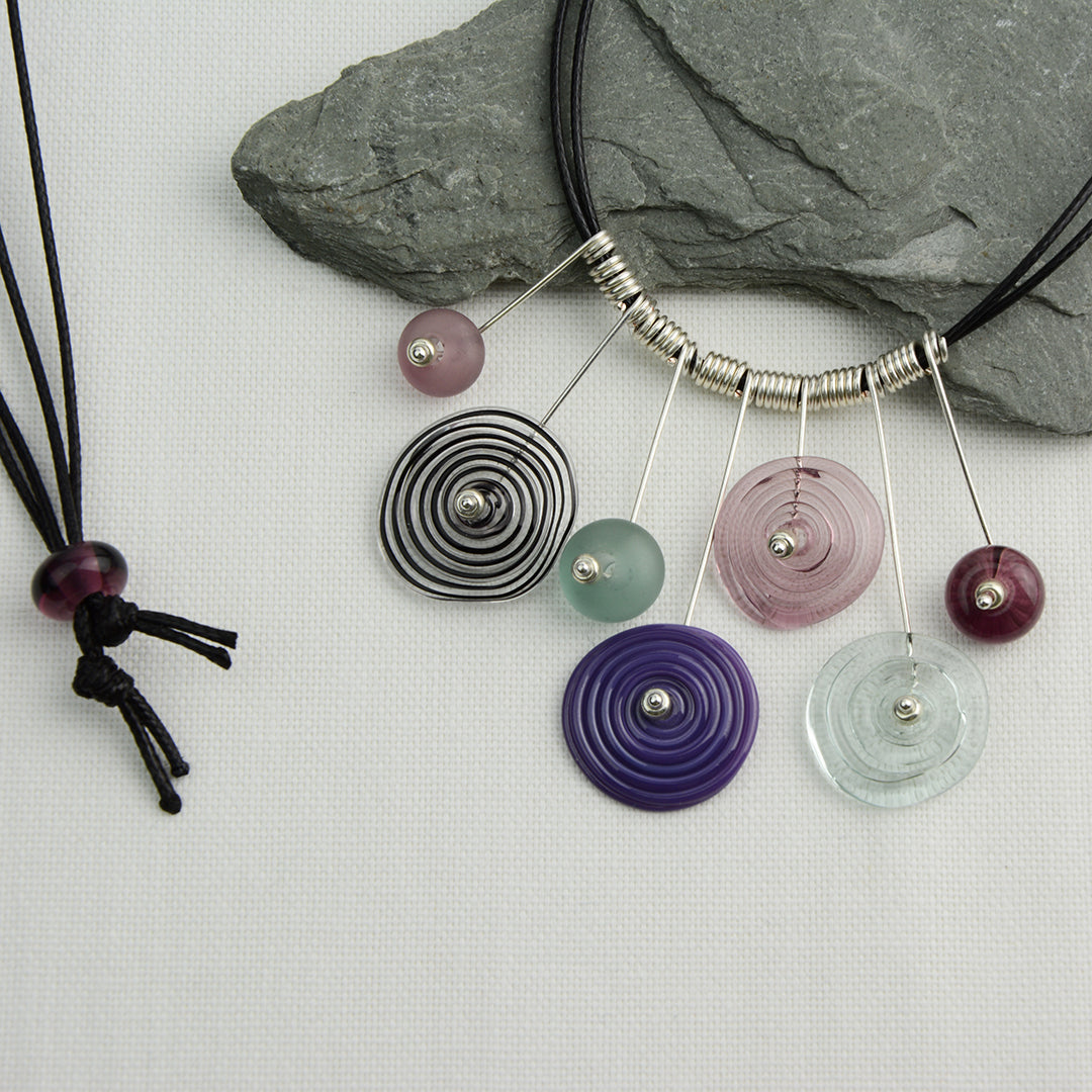 Four spiral beads and three spacer beads in shades of purple and grey on silver headpin drops with silver spiral spacers on black necklace cord