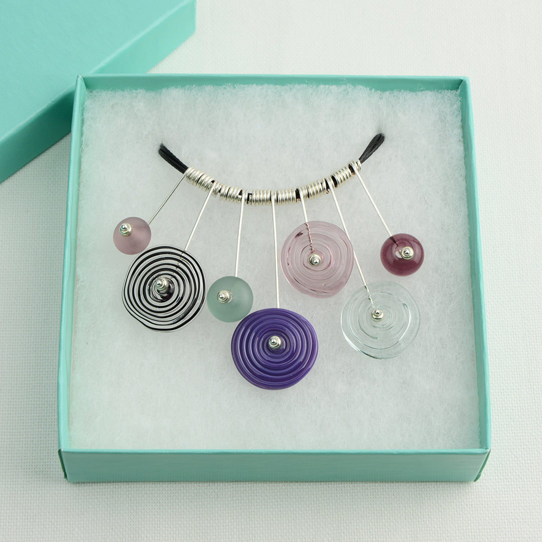 Four spiral beads and three spacer beads in shades of purple and grey on silver headpin drops with silver spiral spacers on black necklace cord in turquoise gift box