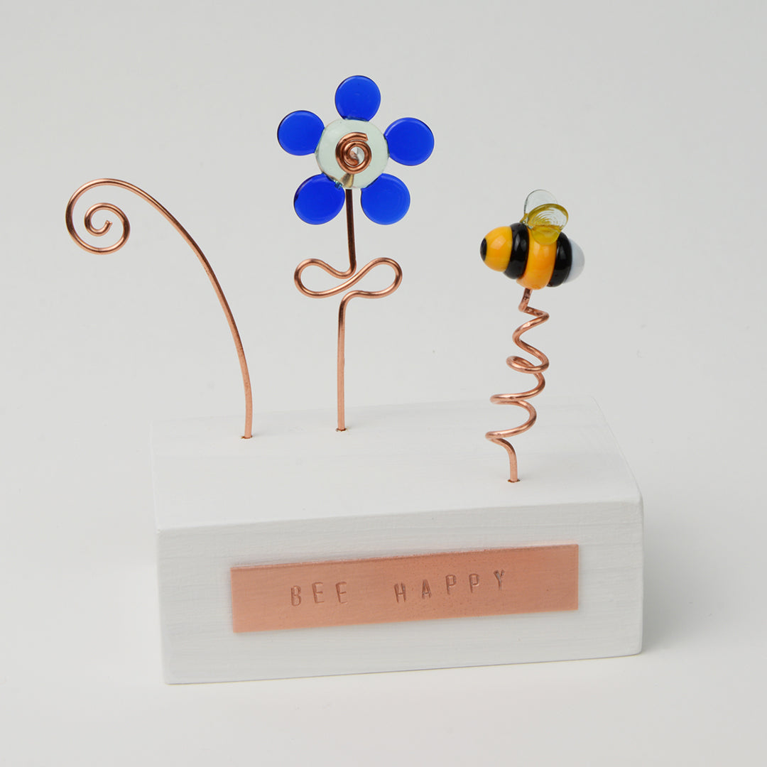 White wooden block with blue glass flower and bee with copper plaque reading Bee Happy