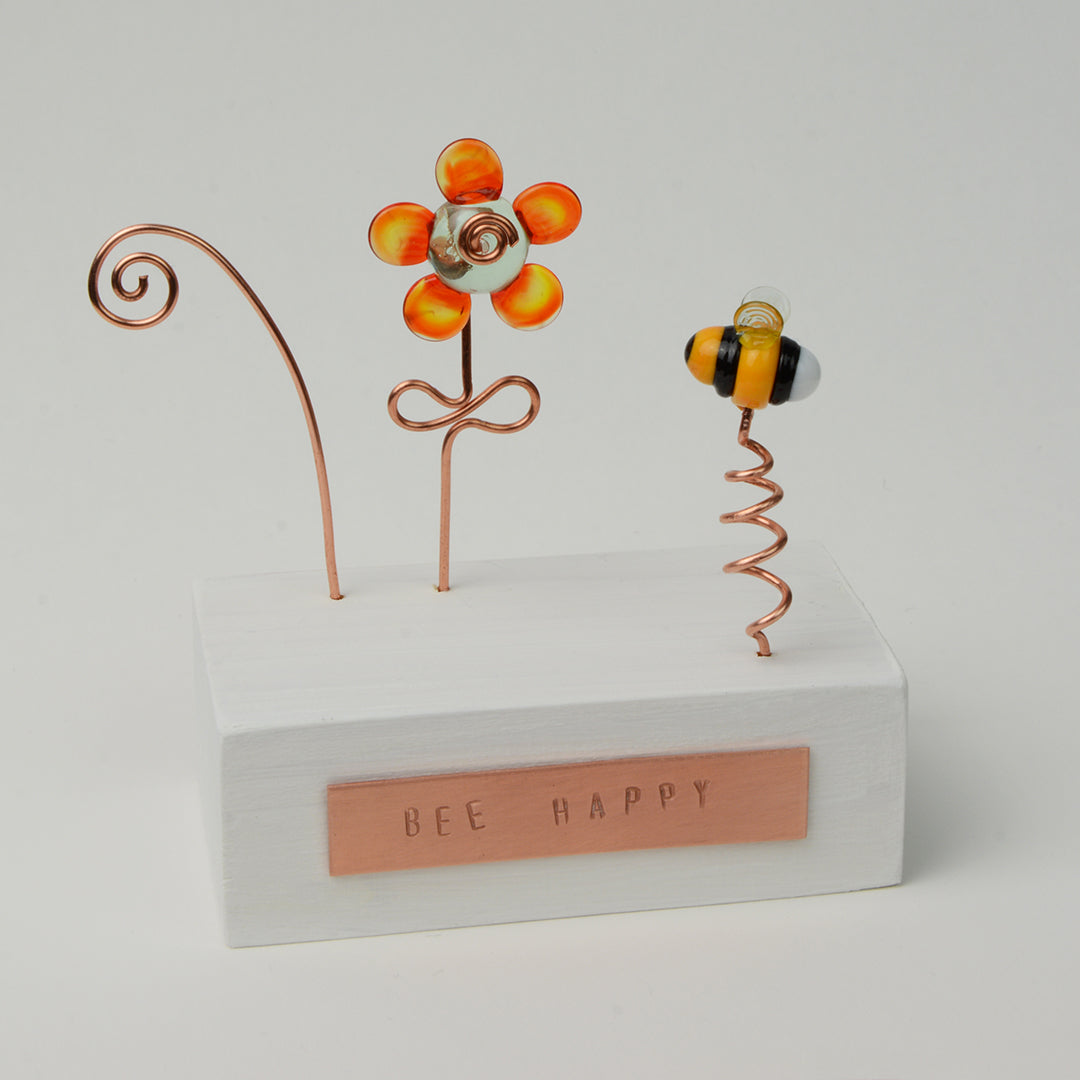 White wooden block with orange glass flower and bee with copper plaque reading Bee Happy