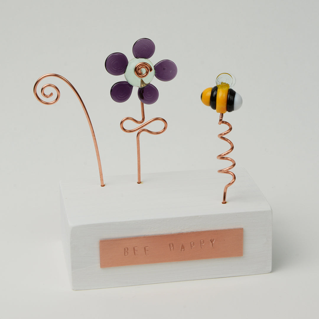 White wooden block with purple glass flower and bee with copper plaque reading Bee Happy