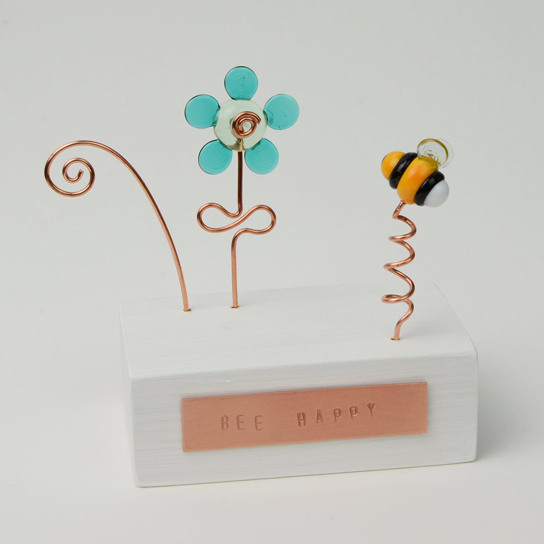 White wooden block with teal glass flower and bee with copper plaque reading Bee Happy