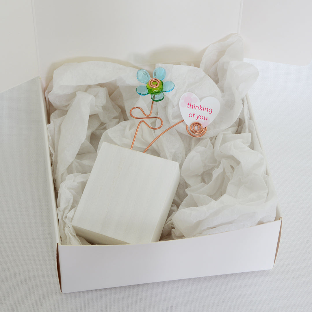 Turquoise and green glass flower  on a white wooden block with a paper heart with the message 'Thinking of You' placed on tissue paper in a white gift box