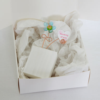 Turquoise and green glass flower  on a white wooden block with a paper heart with the message 'Thinking of You' placed on tissue paper in a white gift box