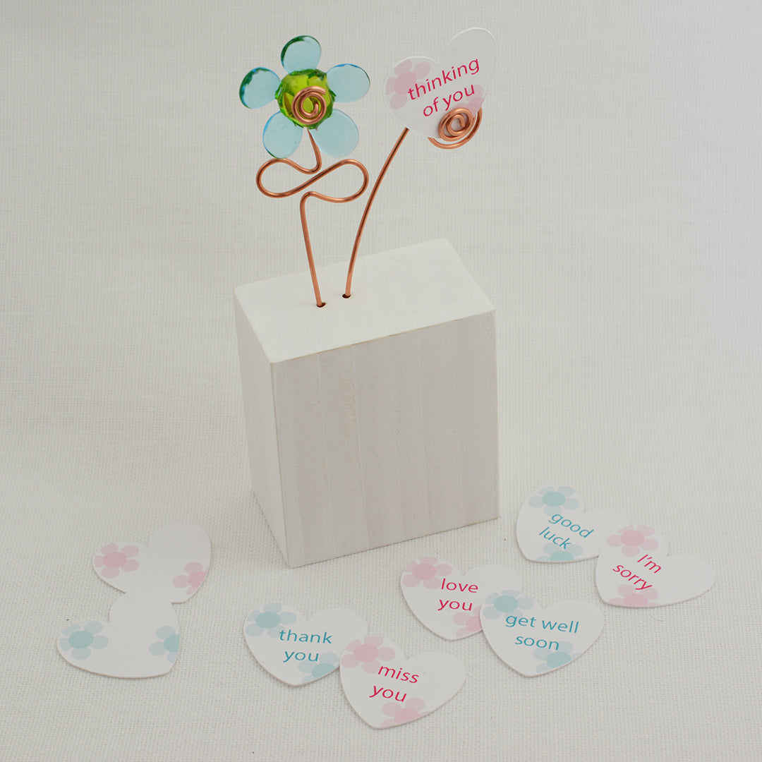 Turquoise and green glass flower  on a white wooden block with a paper heart with the message 'Thinking of You' and other paper hearts showing different messages