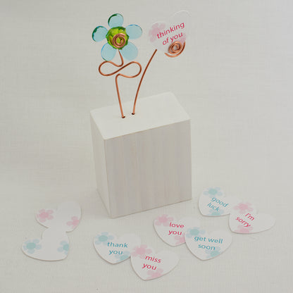 Turquoise and green glass flower  on a white wooden block with a paper heart with the message 'Thinking of You' and other paper hearts showing different messages