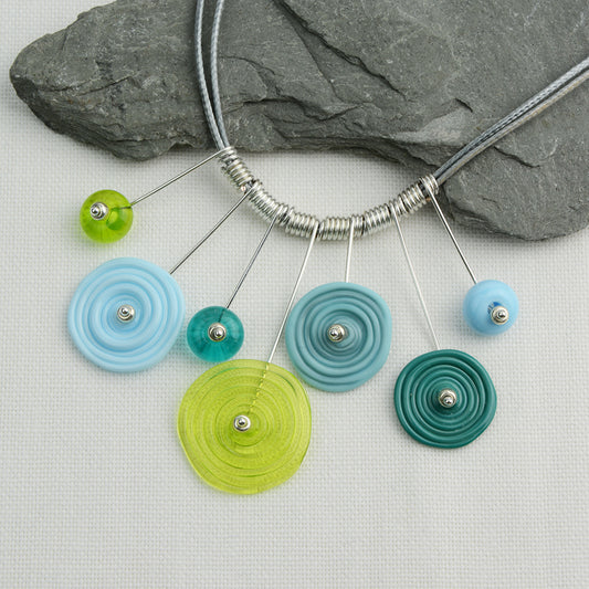 Four spiral beads and three spacer beads in shades of blue and green on silver headpin drops with silver spiral spacers on grey necklace cord